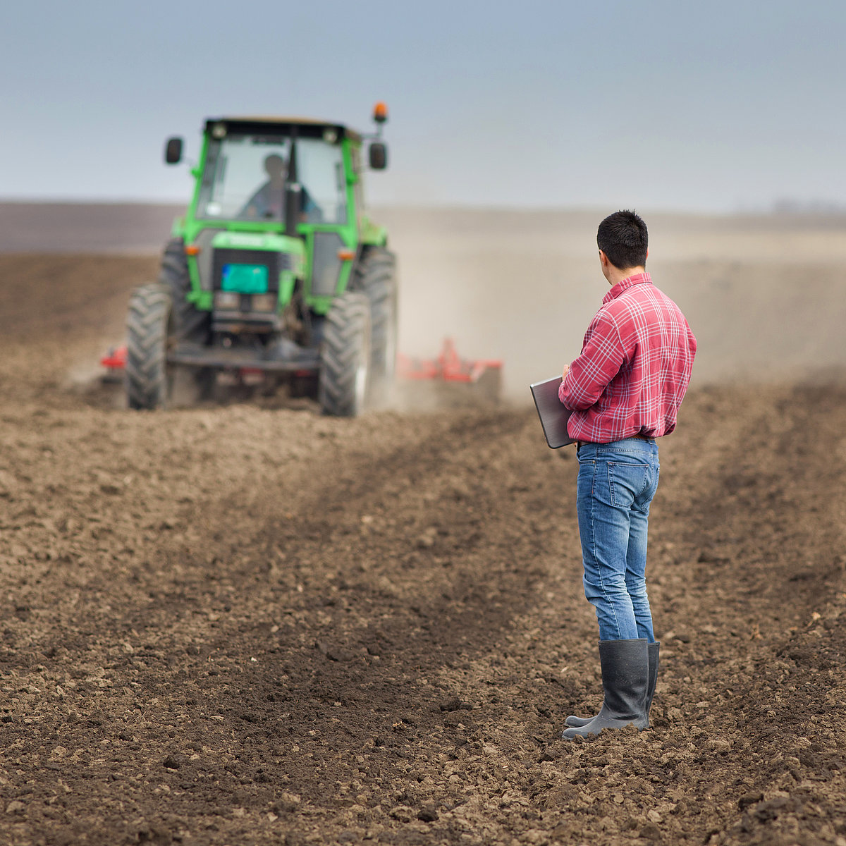 Mann steht mit Tablet auf dem Feld, Traktor fährt im Hintergrund